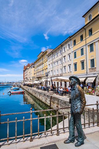 People relaxing on the banks of the Canal Grande of Trieste, a large navigable canal built in 1754-1756 in the center of the city. The statue of James Joyce stands on the Ponte Rosso since 2004, created by the Trieste sculptor Nino Spagnoli on the centenary of Joyce's arrival in Trieste.