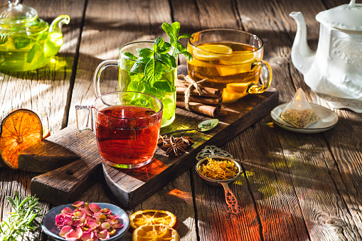 Tea and herbal infusion with dried herbs on wooden rustic table board, including calendula, rose petals, mint leaves, cinnamon sticks, dried orange slices, with mint tea, ginger lemon tea and black tea.