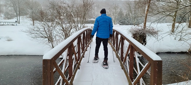 Guy in snow gear is snowshoeing across a snow covered bridge in a snowstorm; active adults, healthy lifestyle, stay fit in the pandemic ideas.