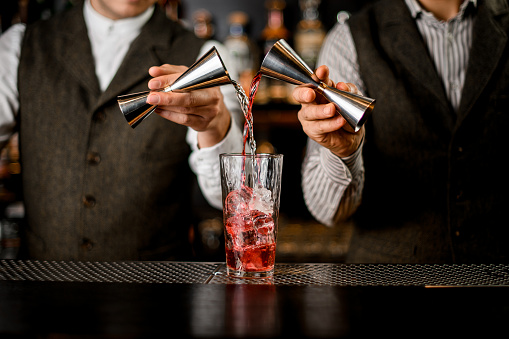Two bartenders hold steel jiggers in their hands and accurate pour drink into transparent glass with ice cubes