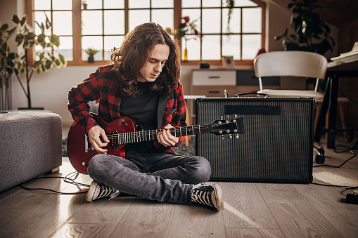 One man, teenage male guitarist with long hair playing electric guitar in living room on the floor at home.