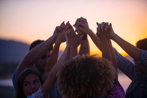 Group of people holding hands in celebration and friendship. Multi ethnic and multi cultural group of young people holding their hands high outdoors on the beach. Shot is a silhouette at sunset or sunrise