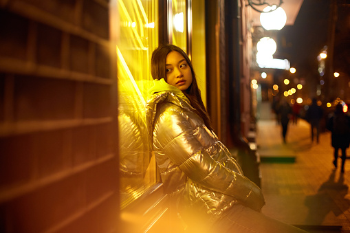 Night illumination concept. Dreamy teenager hipster girl sitting near showcase window with glowing neon light, looking aside