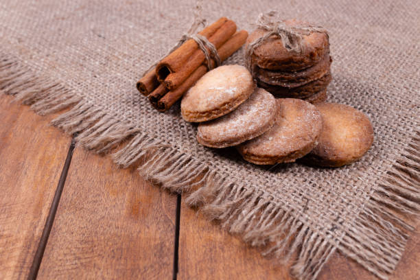side view at an angle of a stack of cookies tied with a string, cinnamon sticks tied with a string, four cookies lying on top of each other, all this lies on a gray rug edged with fringe, and under it is a wooden surface vista lateral en un ángulo de una pila de galletas atadas con una cuerda, palos de canela atados con una cuerda, cuatro galletas que se encuentran una encima de la otra, todo esto se encuentra en una alfombra gris bordeada con flecos, y debajo de ella hay