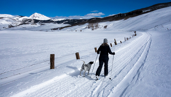 A woman enjoys cross country skiing, with her dog, along a beautiful mountain road.