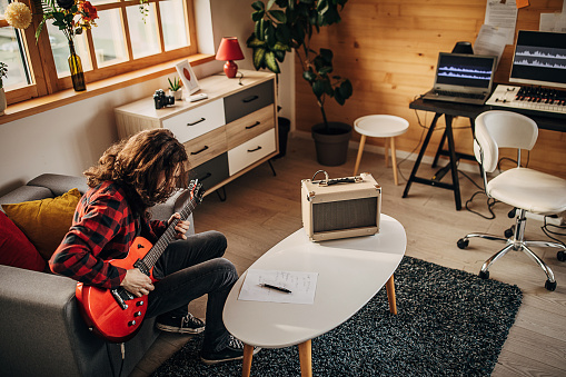 One man, teenage male guitarist with long hair playing electric guitar in living room at home.