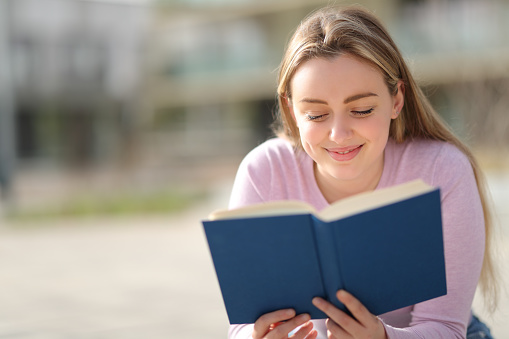 Cheerful caucasian student guy reading book outdoors, preparing for lectures while lying on lawn in university campus or park, free space