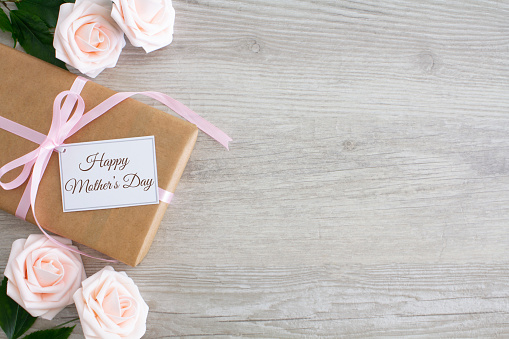 Pale pink roses and gift tied with pink ribbon and gift tag saying Happy Mother's day, shot from above on pale rustic wood.\nCopy space to the right of the image. \nMother's Day flat lay.