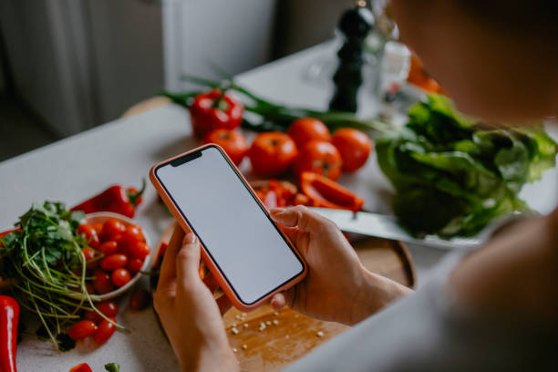 Home Cooking Classes: Hands of a Woman Holding a Mobile Phone Above a Cutting Board Full of Vegetables Anonymous woman following a live cooking session on her mobile phone while making a delicious lunch. people holding board stock pictures, royalty-free photos & images