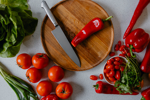 Kitchen knife, chopped red pepper, and half pepper on cutting board. Garlic and spoon with tomato sauce. Onion, tomato and gray napkin on table. Flat lay. Grey background.
