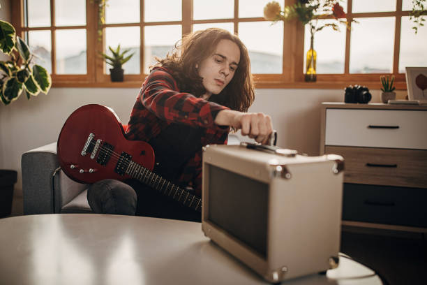 One man, teenage male guitarist with long hair making music on electric guitar in living room at home, adjusting volume on amplifier.