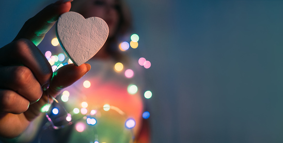 Love symbol. Romantic background. Festive decoration. Woman holding ceramic heart with fingers in colorful neon bokeh lights blur rainbow glow isolated on navy blue copy space.