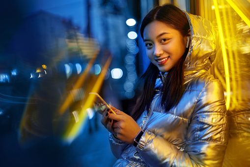 Young stylish asian woman wwearing glossy jacket and wireless earphones standing on the street in fluorescent neon yellow lights listening to music holding smartphone browsing playlist looking camera smiling cheerful