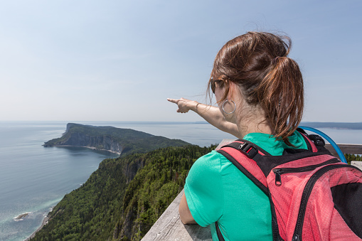 Woman hiker wearing a backpack on a Mountain Summit and Pointing a Direction and the ocean. She is pointing the Gaspe Peninsula in Forillon National Park, Quebec, Canada.
