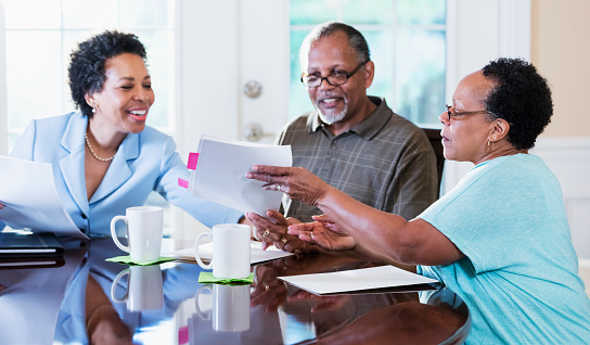 A senior African-American couple in their 60s sitting at home at their dining room table, looking at documents and talking with a financial advisor. The advisor is a mature African-American woman in her 50s.
