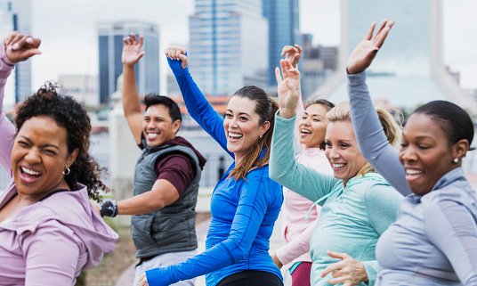 A multi-ethnic group of five women and a man in a city park taking a dance exercise class. They are in their 30s and 40s. The focus is on the mid adult Hispanic woman in the middle.