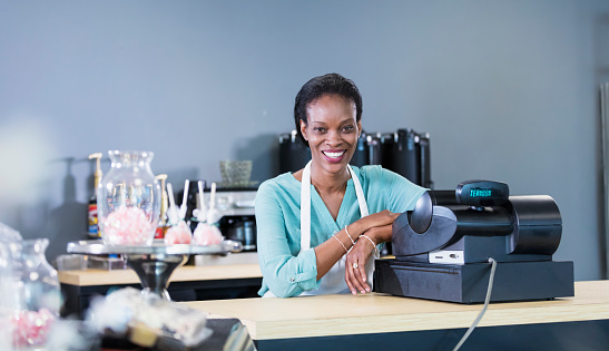 An African-American woman working in a coffee shop, standing at the checkout counter, smiling at the camera, cake pops out of focus in the foreground.