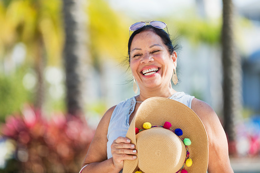 A senior Hispanic woman in her 60s standing outdoors in front of a swimming pool, holding a wide brimmed hat, laughing and looking at the camera.
