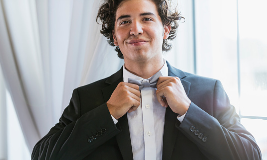 A mixed race teenage boy, 16 years old, standing by a window, wearing a tuxedo. He is dressed for prom. Or perhaps he is the best man at in a wedding. He is fixing his bow tie.