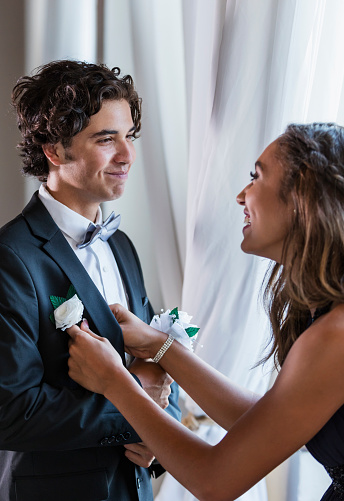 A teenage couple dressed for prom, standing by a window. They are 16 years old. The boy is mixed race Hispanic, Native American and Caucasian. His girlfriend is African-American and Caucasian. She is helping him with his boutonniere.