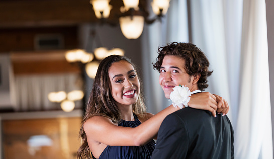 A teenage couple dressed for prom, slow dancing by a window. They are 16 years old. The boy is mixed race Hispanic, Native American and Caucasian. His girlfriend is African-American and Caucasian.  They are looking at the camera.