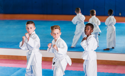 A multi-ethnic group of three boys in a taekwondo class. They are standing side by side with serious expressions, fists raised, wearing training uniforms with white belts. The children are 7 years old. The one on the left is mixed race Hispanic and Caucasian.