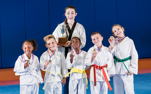 A multi-ethnic group of five children wearing taekwondo uniforms proudly showing off their medals for winning a competition, and their coach is holding the trophy. The boys are 7 years old. The African-American girl is 6 and the other girl is 9.