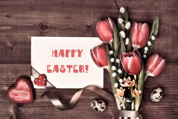 Card with text Overhead view, flat lay. Arrangement of Easter, springtime decorations. Wrapped gifts, red tulips, pussy willow, lily of valley flowers. Easter eggs, hearts, ribbons on brown wood.