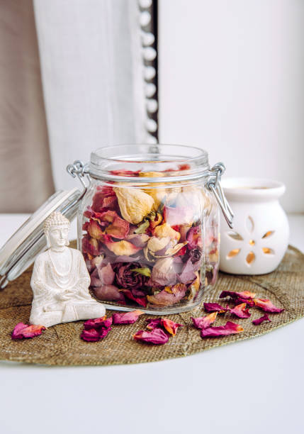 Using dry rose petals to make rose potpourri which is great for home scent. Mason jar filled with dry rose petals and puds with meditating Buddha figurine and aroma lamp with candle next to it.