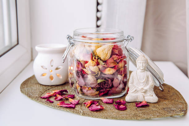 Using dry rose petals to make rose potpourri which is great for home scent. Mason jar filled with dry rose petals and puds with meditating Buddha figurine and aroma lamp with candle next to it.