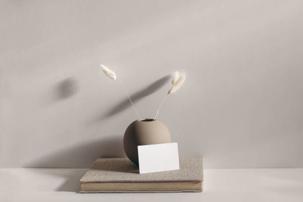 istock Modern stationery still life scene. Beige spheric vase with dry lagurus grass. Table background in sunlight. Blank business card mockups lean on old book. Champagne wall, long shadows. 1301165737