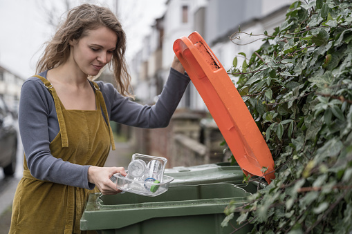 Partial side view of longhaired British woman in casual clothing lifting lid of garbage bin in front of London home to recycle plastic containers.