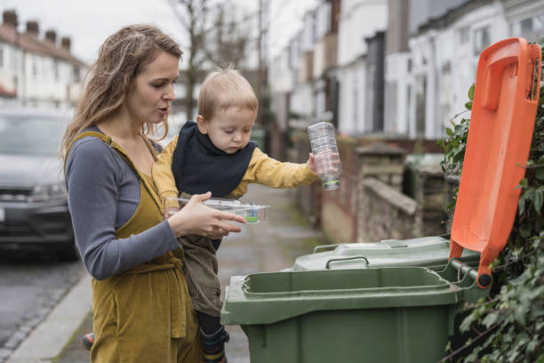 Responsible British mother teaching toddler how to recycle Partial side view of longhaired Caucasian woman in casual clothing holding 2 year old son in her arms as he puts plastic containers in recycling bin. social responsibility photos stock pictures, royalty-free photos & images