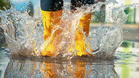 LOW ANGLE, CLOSE UP, DOF: Unrecognizable Female in brand new bright yellow rubber boots jumps into the glassy puddle on the sidewalk. Carefree young woman in rain boots jumps into the big puddle.