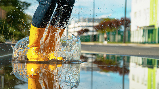 CLOSE UP, LOW ANGLE, DOF: Female in bright yellow rubber boots jumps into the glassy puddle on the side of the empty road. Crystal clear water splashes as unrecognizable woman jumps into a puddle.