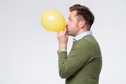 Young man blowing a yellow balloon training his lungs after coronavirus. Studio shot