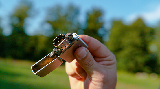 CLOSE UP, DOF: Unrecognizable young man skilfully flicks his metal lighter open during a fun summer picnic. Unknown male camper pops the top off a vintage silver lighter to light up a campfire.