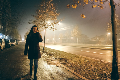 Young woman is using phone, scared of walking down the dark street and surrounded with fog.