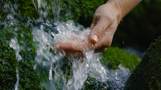 CLOSE UP, DOF: Thirsty person uses their hand to scoop up a handful of refreshing cold stream water. Unrecognizable woman outstretches hand into a cascading stream of crystal clear river water.