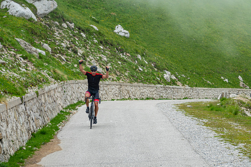 Athletic man victoriously lifts his arms while cycling up a mountain road in the misty Julian Alps. Excited male tourist road cycling up Mangart celebrates finishing a challenging climb on foggy day.