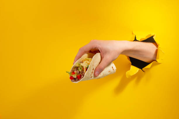Hand giving a taco through a torn hole in yellow paper background Hand giving a taco through a torn hole in yellow paper background. Tasty mix of vegetables, meat and cheese in a tortilla. tacos stock pictures, royalty-free photos & images