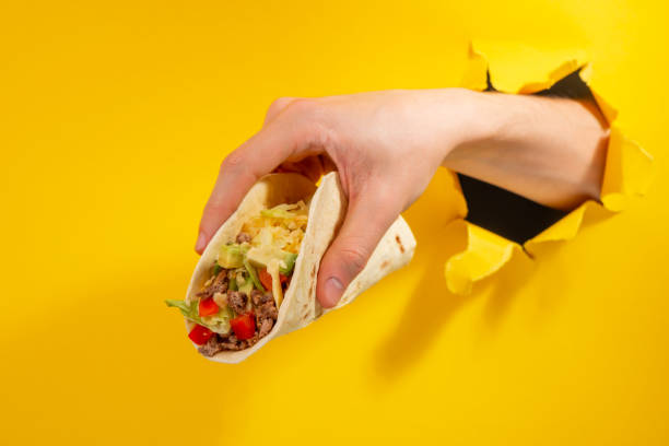 istock Hand holding a taco through a torn hole in yellow paper background 1301180015