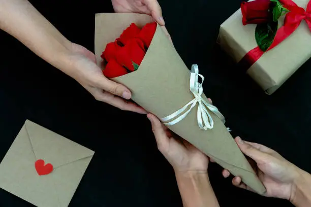 Photo of Close-up of Women giving a bouquet of roses with the gift box, card letter with red heart on the table, black background. Concept of creating handmade gifts for Valentine's Day or other days holiday