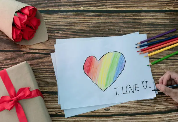 Photo of Woman handwriting word I LOVE U and heart with rainbow flag as a symbol of LGBT on white paper with colored pencils, gift box, bouquet of roses. Concept of creating handmade gifts for Valentine's Day