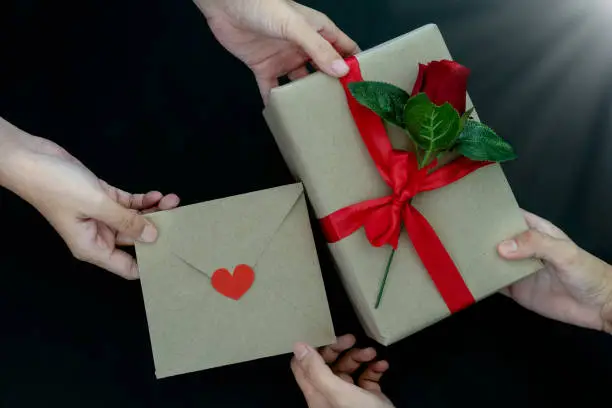 Photo of Two Women hands changing presents and celebrating holidays together. Romantic couple giving gift box and a red rose, letter with red heart, as handmade gifts for Valentine's Day or other days holiday