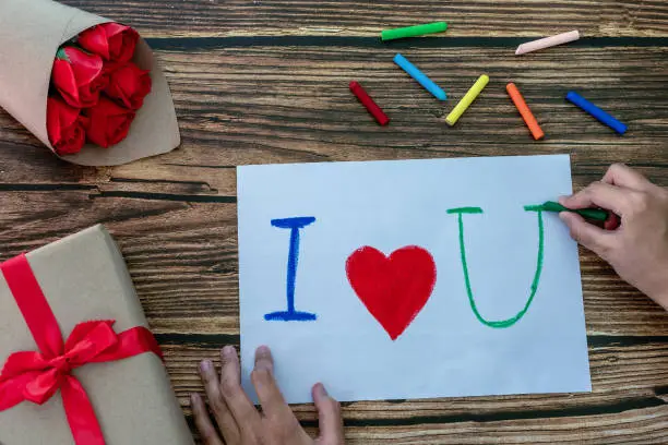 Photo of Woman handwriting words I and U and red heart on middle white paper with colorful crayons, gift box and bouquet of roses on the wooden table. Concept of creating handmade gifts for Valentine's Day