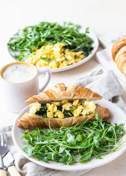istock Delicious croissant sandwich with scrambled spinach eggs served with arugula on plate and coffee cup, light concrete background. 1301198295