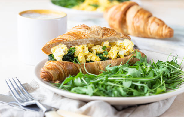 istock Delicious croissant sandwich with scrambled spinach eggs served with arugula on plate and coffee cup, light concrete background. 1301198341