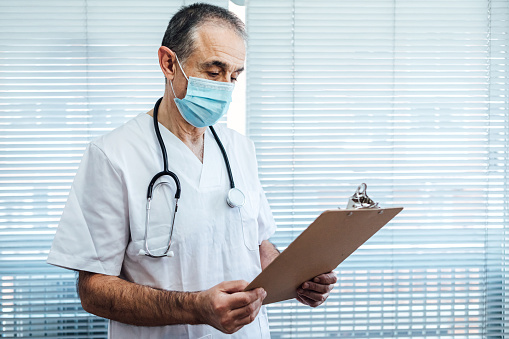 Mature male doctor - nurse wearing face mask, talking on mobile phone and holding a report next to a hospital window. Covid-19 and medicine concept