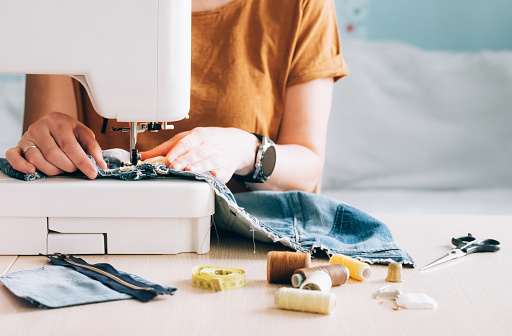 A woman tailor works at a sewing machine sews reuses fabric from old denim clothes. The concept of economical reuse of recyclable things. Homemade needlework hobby. Selective focus